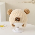 Close-up of beige knitted baby hat with dual pom-poms and cartoon dinosaur patch detail - Dinosaur Label - Beige