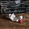 Resin skull figurine reading a red book on a dark wood table for Halloween decor - Skeleton Lying Reading A556-0006