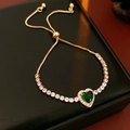 Close-up of golden adjustable heart bracelet with green crystal on black leather surface - Gold-Green