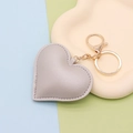 Détail macro d'un porte-clés cœur en similicuir gris sur un plateau beige avec anneau doré - Fermoir Chaîne Dorée / Argenté
