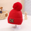Close-up of a red knitted kids hat with a fluffy pom-pom and a stylish OK letter patch - One Size (2-5 Years) / Red