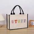 Close-up of colorful embroidered STUFF lettering on beige canvas tote bag with black handles - STUFF