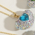 Dettaglio macro di un ciondolo a cuore dorato con gemma blu e cristalli colorati - Zirconia Blu (XL2601-3)