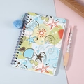 Primer plano de un cuaderno de espiral con tema marino y un colgante de pompón azul - Mundo Marino