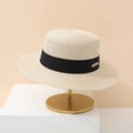 Close-up of beige woven straw boater hat featuring a black ribbon and gold logo plaque - Beige