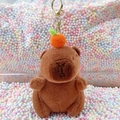 Brown plush capybara keychain with orange hat charm on colorful foam background - Dark Brown Orange + Gold Clip