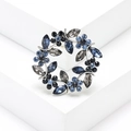 Elegant olive branch wreath brooch with blue and grey crystals on a white pedestal - Blue