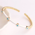 Detailed shot of gold plated evil eye cuff bracelet with white enamel on ceramic tray - White