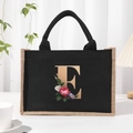 Black linen tote bag featuring a gold letter E and floral print with braided handles - Bronze Floral Letter E