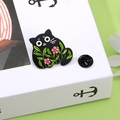 Spilla smaltata gatto nero a fumetti con fiori rosa e foglie verdi su sfondo bianco - Gatto Nero Con Fiore Rosa E Foglie Verdi