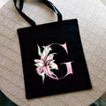 Black canvas tote bag with pink letter G and lily print on a textured background - Letter G / Black