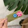 Detail of green plush hair clip with blue button and pink bow accents held by hand - Green
