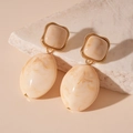 Detail van beige hars oorbellen met marmerpatroon en gouden rand op een stenen vlak - Broken Wit