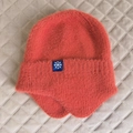 Dettaglio del berretto invernale rosso corallo con toppa a fiocco e paraorecchie in maglia - Taglia unica / Rosso Mattone