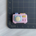 Pink resin cartoon camera charm with blue lens detail on a grid cutting mat - Inflatable Radio Prop