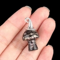 Dark mottled stone mushroom pendant with silver wizard hat detail held by fingers - Gun Black Cap / Rhodonite