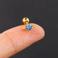 Macro shot of gold heart arrow earring with blue zircon on fingertip for size scale - Gold
