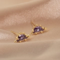 Macro shot of 18K gold dinosaur earrings with purple cubic zirconia on beige silk cloth - Purple