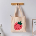 Beige canvas tote bag with red plush strawberry embroidery hanging on a wooden hook - Apricot