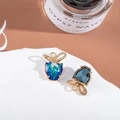Close-up of golden bow and heart crystal earrings displayed on white stone props - Blue Green Gradient/Dark Blue