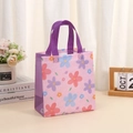 Detail of a pink non-woven gift bag with floral print and purple handles on a desk - Pink Purple Floral / 36x32x12cm