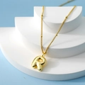 Golden 3D bubble letter R pendant necklace on a geometric display with blue background - Letter R(R)