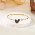 Detail eines goldenen Armreifs mit schwarzem Herzanhänger und funkelndem Zirkonstein - Schwarz