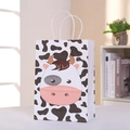 Close-up of a cartoon cow print party gift bag with white braided handles on a table - Cow Pattern (Black & White)