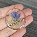 Handmade round resin pendant featuring purple dried flowers and gold leaf in gold frame - Blue Gypsophila