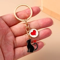 Detail of a golden cat keychain with black enamel feline and red heart charms on a hand - Black