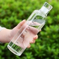 Hand holding transparent glass water bottle with time markers against a green garden - 500ml / Clear