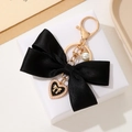 Detalle de llavero con lazo de satén negro, colgante de corazón dorado y perla en caja de regalo - Negro