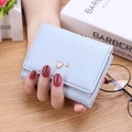 Short tri-fold light blue wallet with gold heart charm held by hand over wooden desk - Light Blue