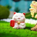 White lucky cat ornament with red fan and gold lettering on green moss background - Money & Leisure Lucky Cat