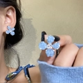 Détail de boucles d'oreilles fleurs bleu clair avec cristaux sur l'oreille et la main - Clous Diamantés Mats (20#)