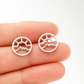 Close-up of circular cutout earrings with sun and mountain pattern held in fingers - Steel