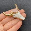Colgante calavera de toro con bordes dorados y gema rosa, sostenido sobre un dedo - N.º 9 - Piedra ovalada rosa claro