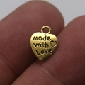 Golden heart-shaped charm with 'made with Love' engraving held on a fingertip - 12*10mm / Antique Gold