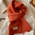 Close-up of soft orange knitted scarf with emo tag on white shirt and beige coat - Pumpkin Orange