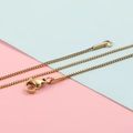 Dettaglio di una collana a catena box dorata con chiusura a moschettone su sfondo bicolore - Placcato Oro 18K