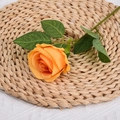 Single orange artificial silk rose with green leaves on a woven mat background - Yellow