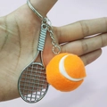 Hand holding a silver tennis racket keychain with a fuzzy orange ball over a pink background - 3.8cm Ball / Orange
