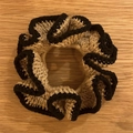 Crochet ruffled scrunchie in beige and black, displayed on a warm wood texture background - Mixed Brown