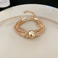 Detail eines mehrlagigen Goldkettenarmbands mit einer großen Metallkugel auf weißem Teller - Armband - Gold