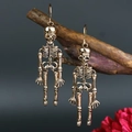 Punk style golden skeleton earrings hanging on a branch against a dark background - Antique Gold
