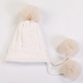 Detail view of white knit hat featuring large faux fur pom-pom and braided hanging cords - White