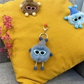 Blue fuzzy plush monster keychain with gray knit hat and glasses shown on a yellow cushion - Haze Blue