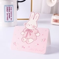 Pink cartoon rabbit greeting card with 'All the best' text in a cozy home setting - Rabbit