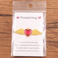 Red enamel heart pocket hug charm with golden wings and emotional card on wood background - Gold Wings Red Heart