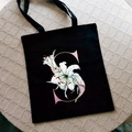 Black canvas tote bag featuring a pink letter S print and white lily flower design - Letter S / Black